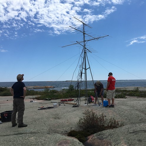 A few people stand looking at a small metal-framed tower (about 18 ft high). They are out on an island on Georgian Bay. It's summer, the sky is clear, and the island has smooth, flat rock.  